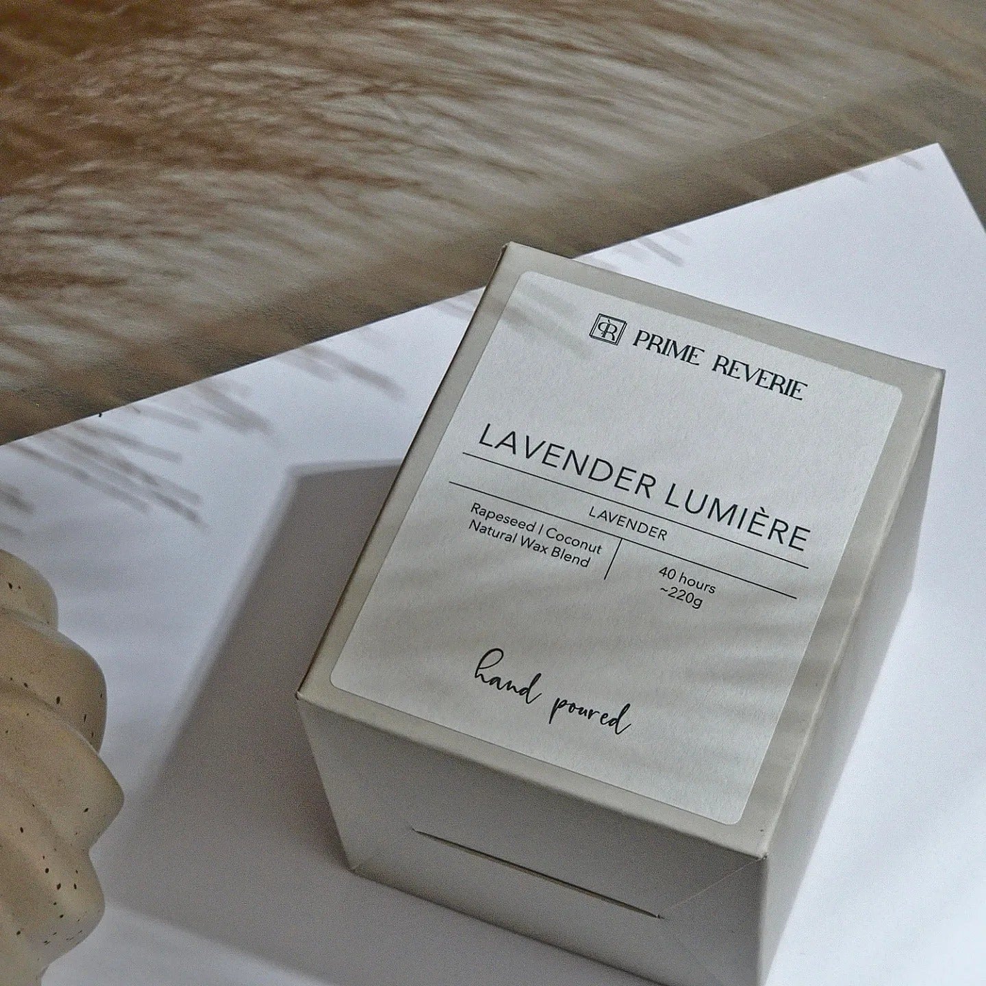 A beige candle labeled "Lavender Lumière" sits on a white surface next to a wavy ceramic object. The label notes a 40-hour burn time and a blend of rapeseed and coconut natural wax.